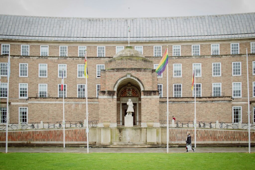 person in black jacket walking near a brown building flags on poles at daytime